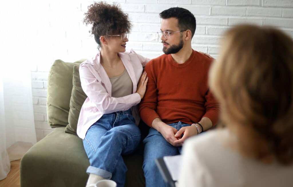 Family Counseling Services: Building Stronger Connections with PS-ITS Counseling 11 A couple sits together on a couch, exchanging looks of understanding and hope, as a therapist from family counseling services listens attentively in the foreground.