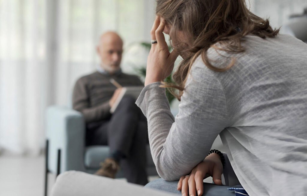 Depression Therapy in Chicago: Compassionate Care for Your Mental Well-Being 1 A person is sitting on a couch with a hand on the forehead, appearing to be in a thoughtful or distressed state. In the background, another person is seated on a chair, holding a notepad and pen, suggesting a therapy session. The setting appears to be a professional environment with soft lighting and a calm atmosphere. This image is relevant as it depicts a scene of depression therapy in Chicago.